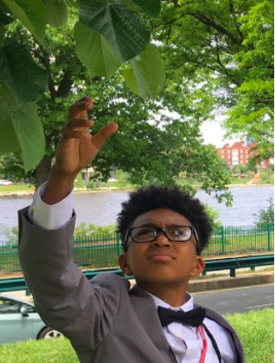Young student, dressed smartly in a suit and bowtie, reaches for a leaf on a tree.